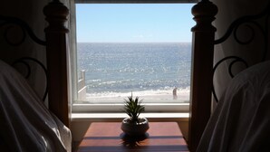Een slaapkamer met uitzicht op de oceaan door een raam, een nachtkastje met een plant en een bed met witte beddengoed.