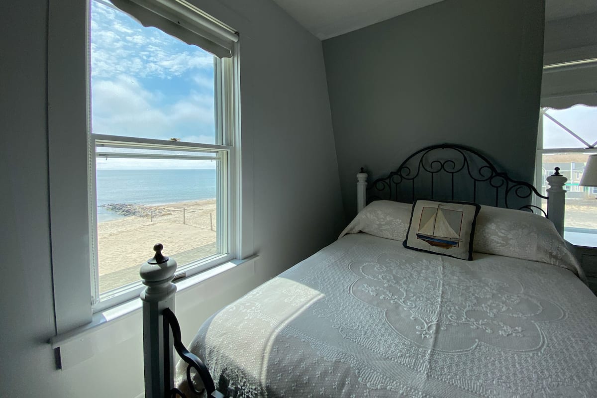 Een slaapkamer met een groot raam dat uitkijkt op een strand, een bed met een geborduurde spreideken en een decoratief kussen.