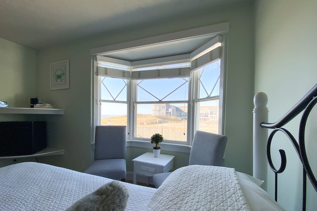 Een slaapkamer met een groot bed, een stoel, een klein tafeltje met een plant en een raam met uitzicht op het strand en huizen.
