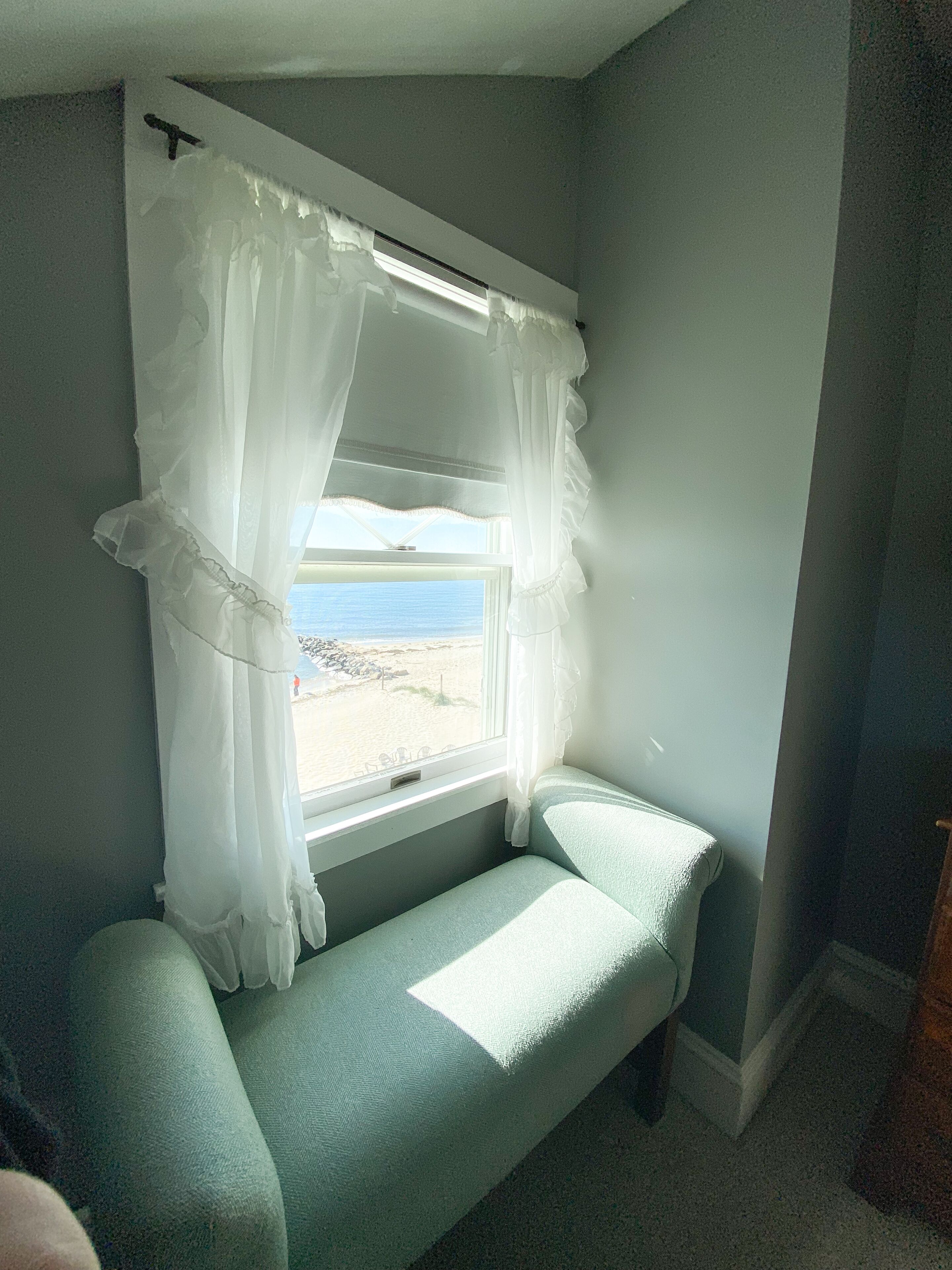 A room with a window overlooking a beach, a small bench, and a view of the sea.