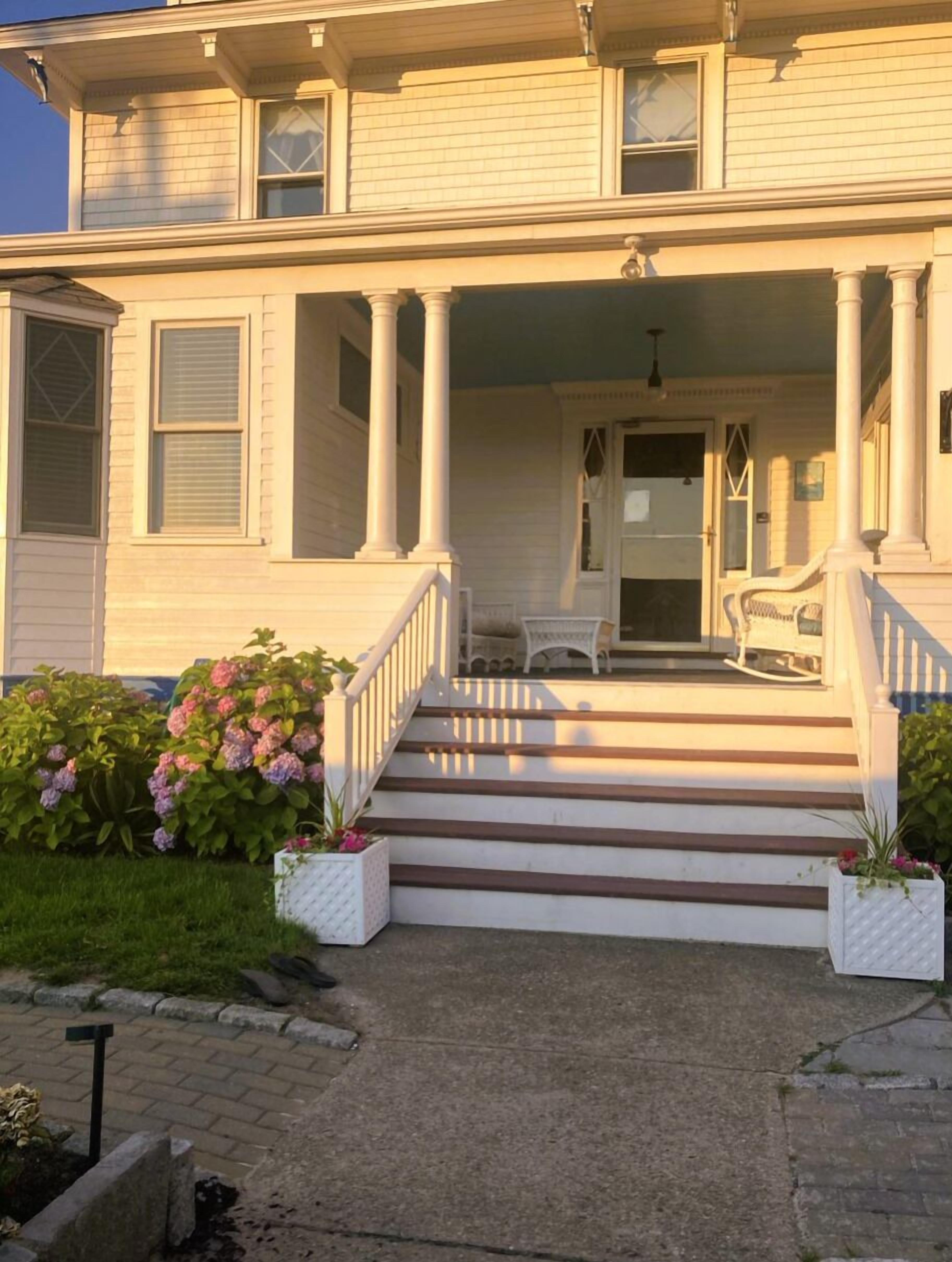 A two-story house with a front porch, white columns, and a small garden with pink flowers.