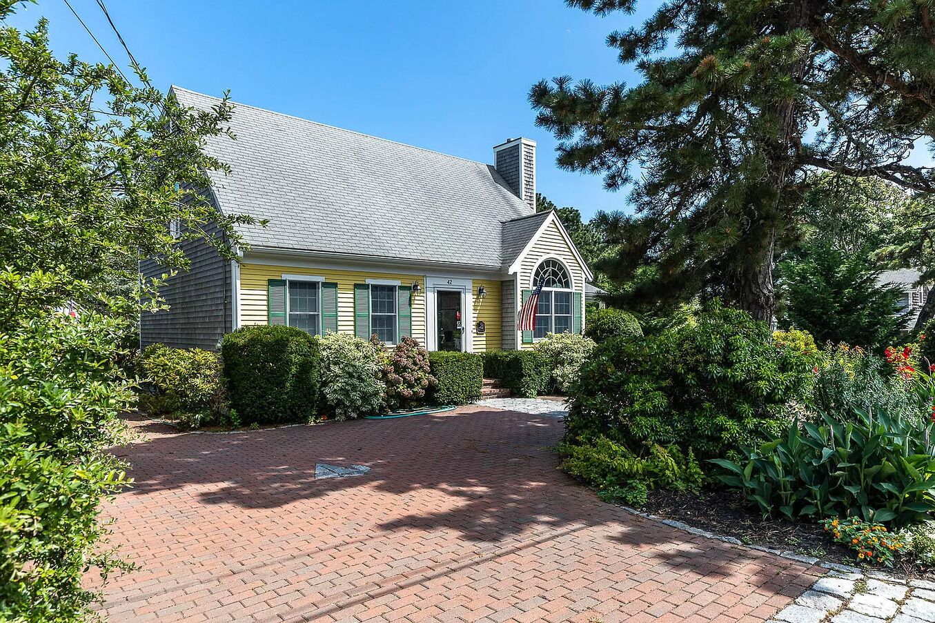 A yellow house with a brick driveway and green shrubs.
