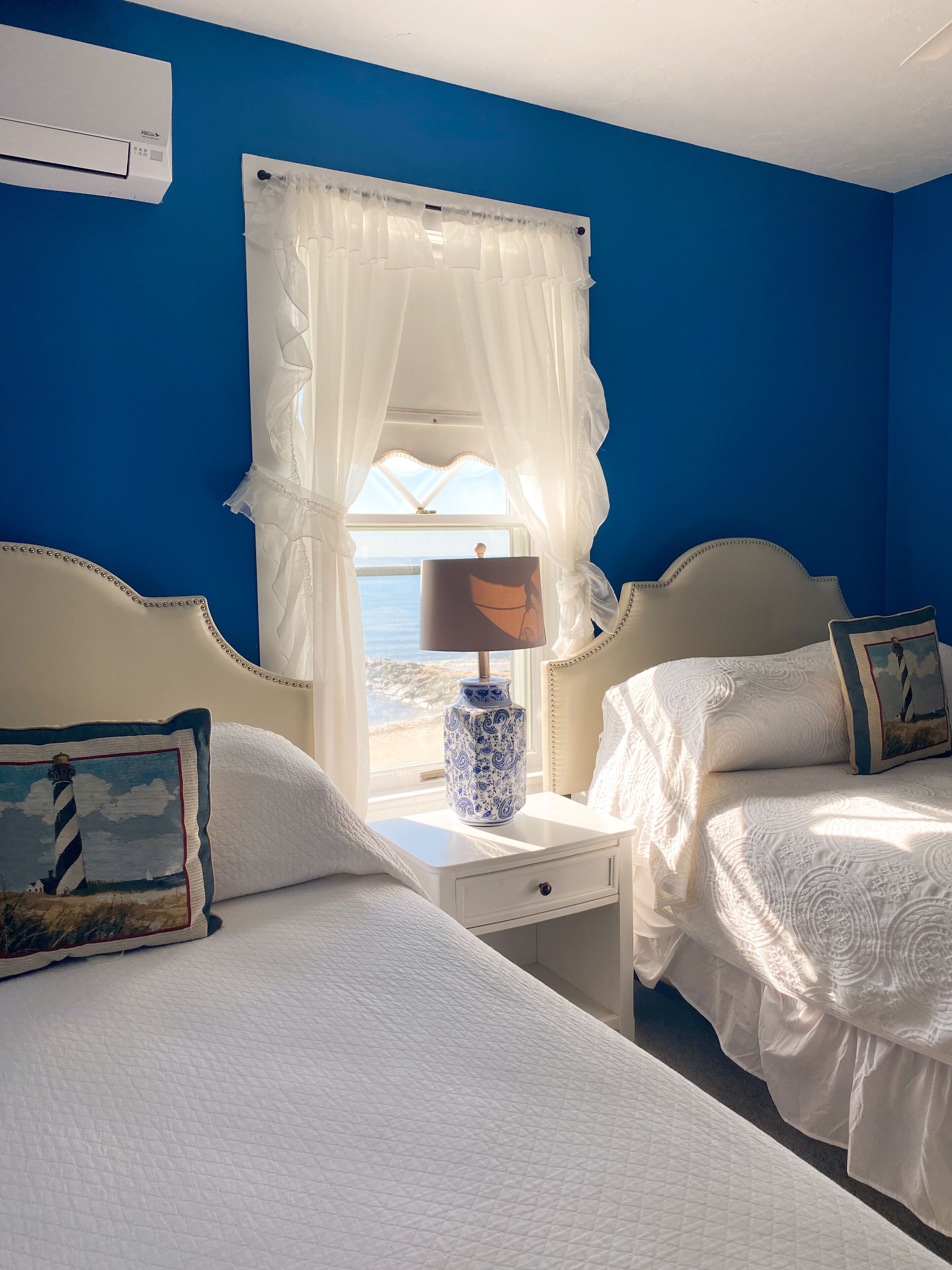 A hotel room with two beds, a nightstand with a lamp, and a view of the sea through sheer curtains.