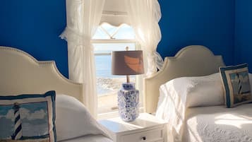 A hotel room with two beds, a nightstand with a lamp, and a view of the sea through sheer curtains.