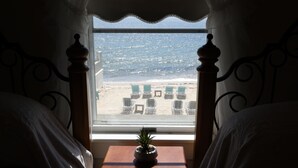 Een slaapkamer met uitzicht op het strand door een raam, een klein tafeltje met een plant en een bed met witte lakens.