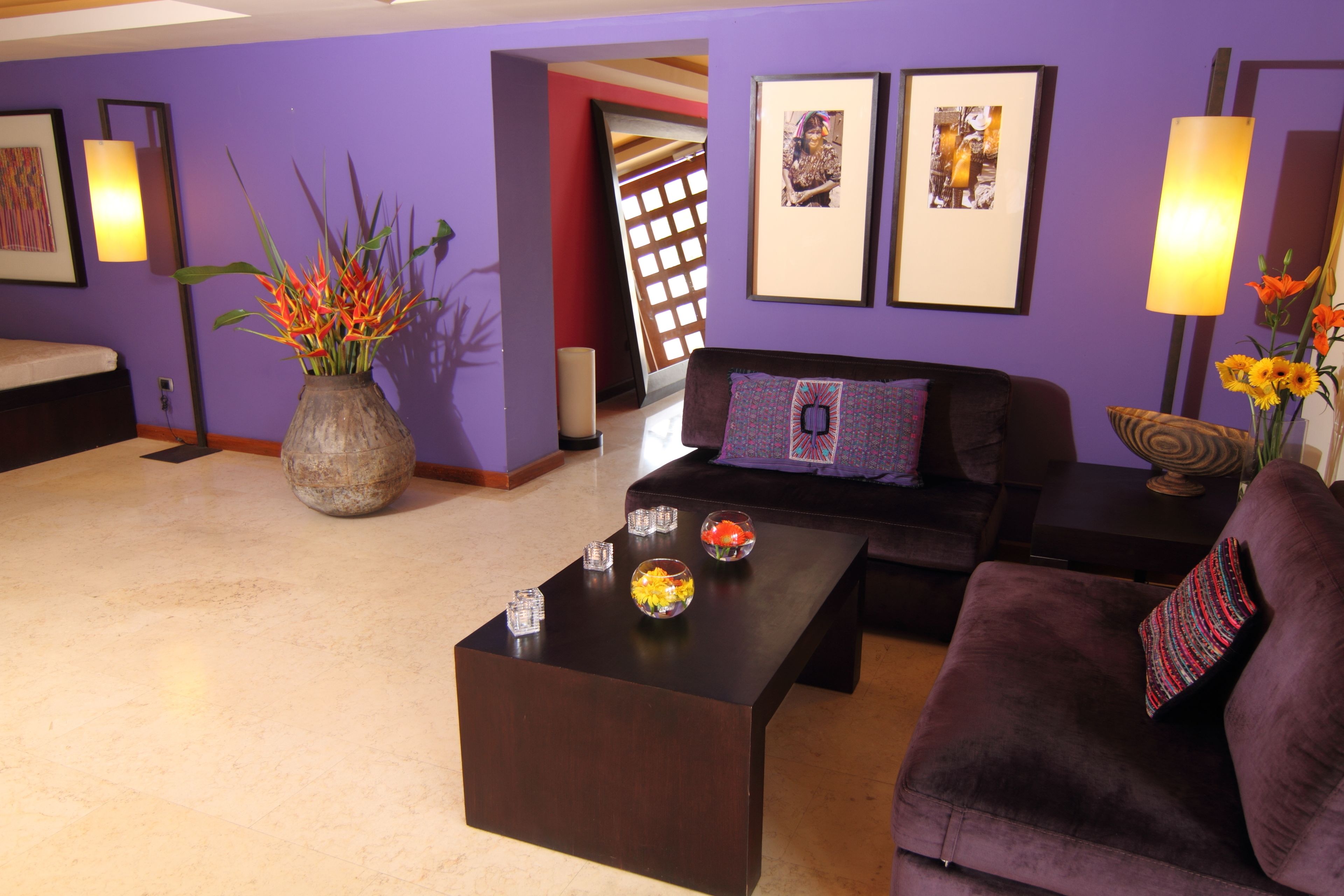 A living room with a dark brown coffee table, a purple sofa, and a large vase with flowers. There are framed pictures on the wall and a lamp on a side table.