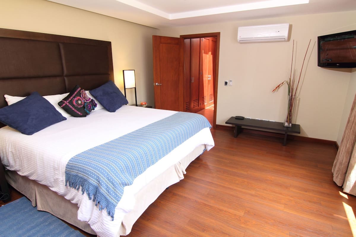 Una habitación de hotel con cama, mesita de noche, lámpara, aire acondicionado, televisor y un banco.