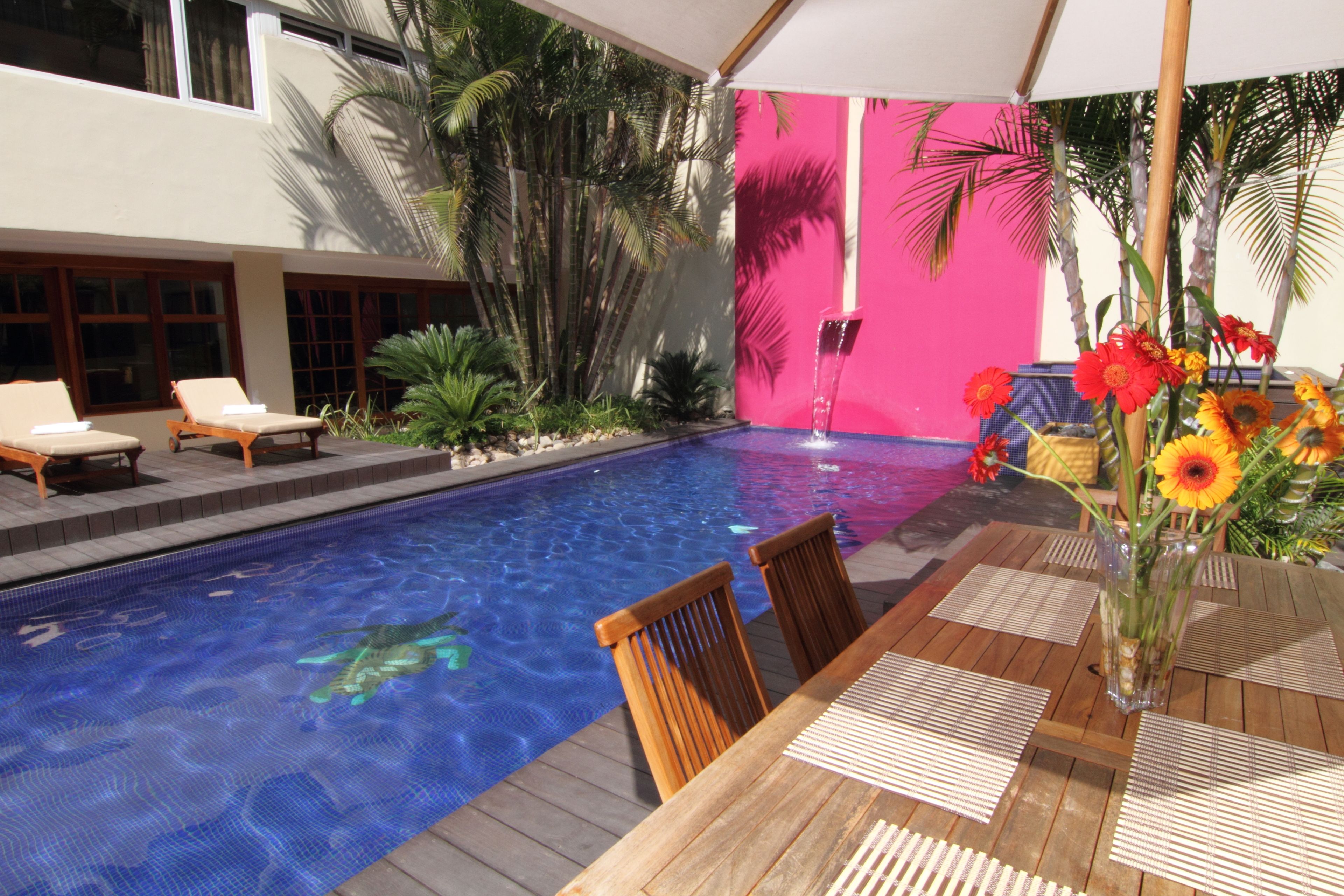A pool area with lounge chairs, a wooden table, and a vibrant pink wall.