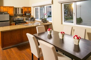 A modern kitchen with wooden cabinets, a stainless steel refrigerator, a microwave, and a stove. There is a dining area with a dark wooden table and four chairs. Two vases with red flowers are on the table.