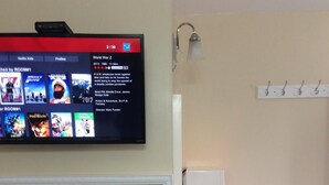 Una habitación con una televisión de pantalla plana mostrando una selección de películas, una pizarra, un escritorio con lámpara y un perchero.