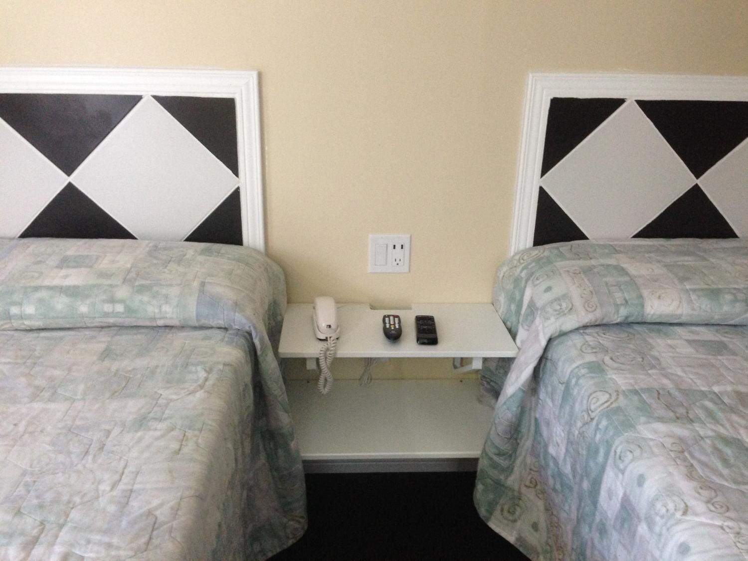 Habitación de hotel con dos camas, una mesita de noche con un teléfono y dos mandos a distancia, y un enchufe en la pared.