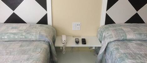 A hotel room with two beds, a bedside table with a phone and two remote controls, and a power outlet on the wall.