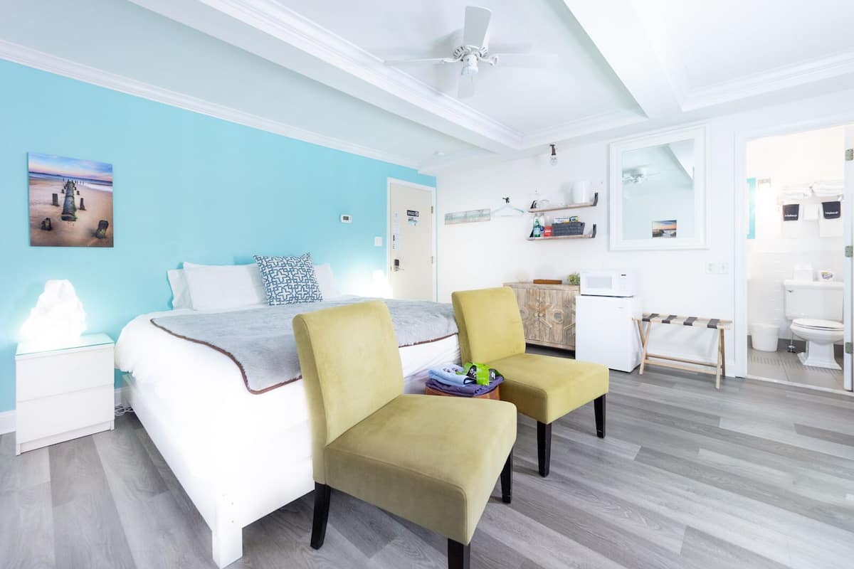 A modern bedroom with a bed, two chairs, a nightstand, a ceiling fan, and a bathroom visible through an open door.
