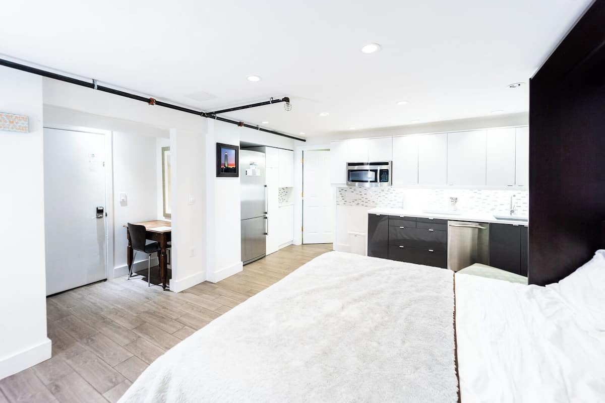 A modern hotel room with a large bed, a desk, a kitchenette, and a bathroom.