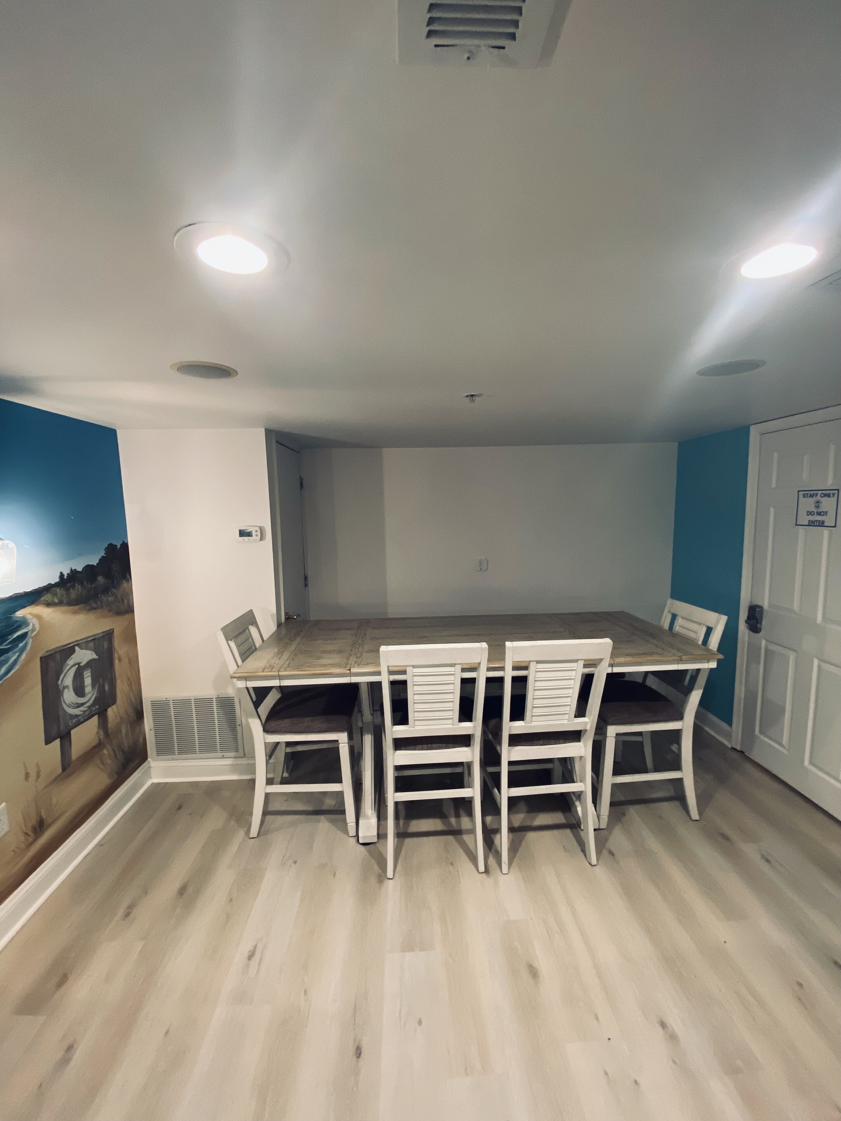 Un comedor con una mesa de madera y sillas blancas, un mural en la pared y una puerta con el letrero 'Staff Only'.