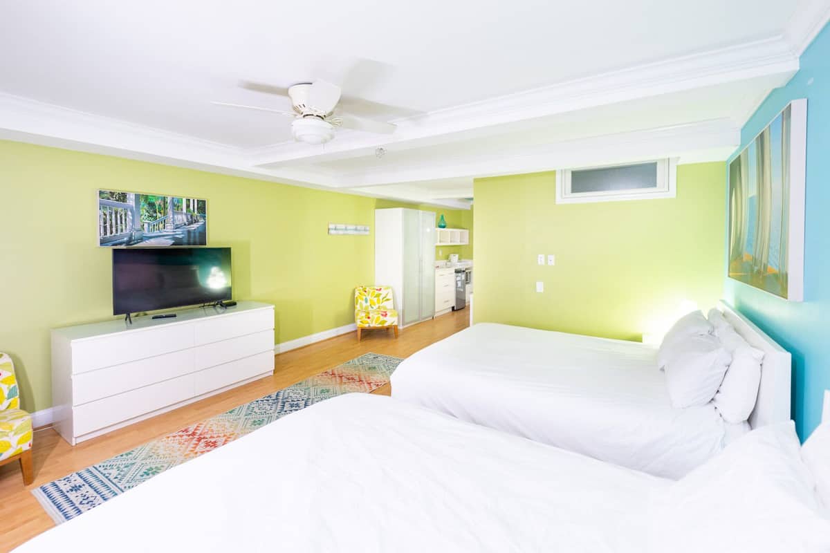 A bedroom with a bed, a TV, a ceiling fan, and a kitchen in the background.
