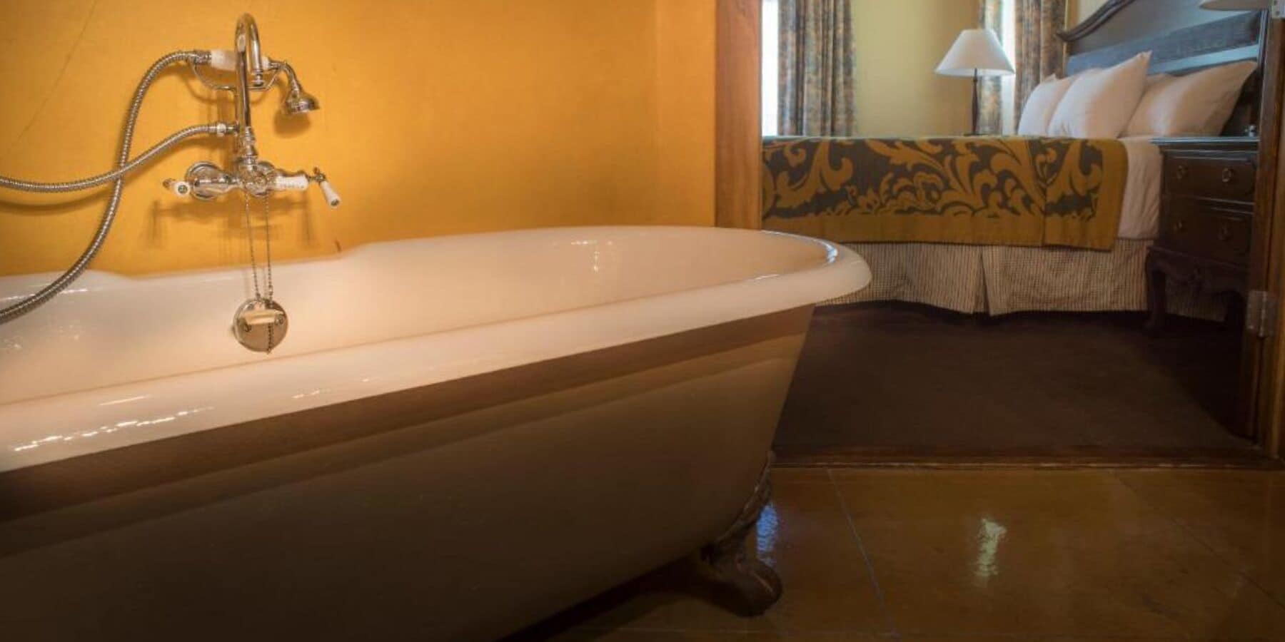 A large bathtub with a shower head, a wooden floor, a bed with a patterned bedspread, and a nightstand with a lamp.