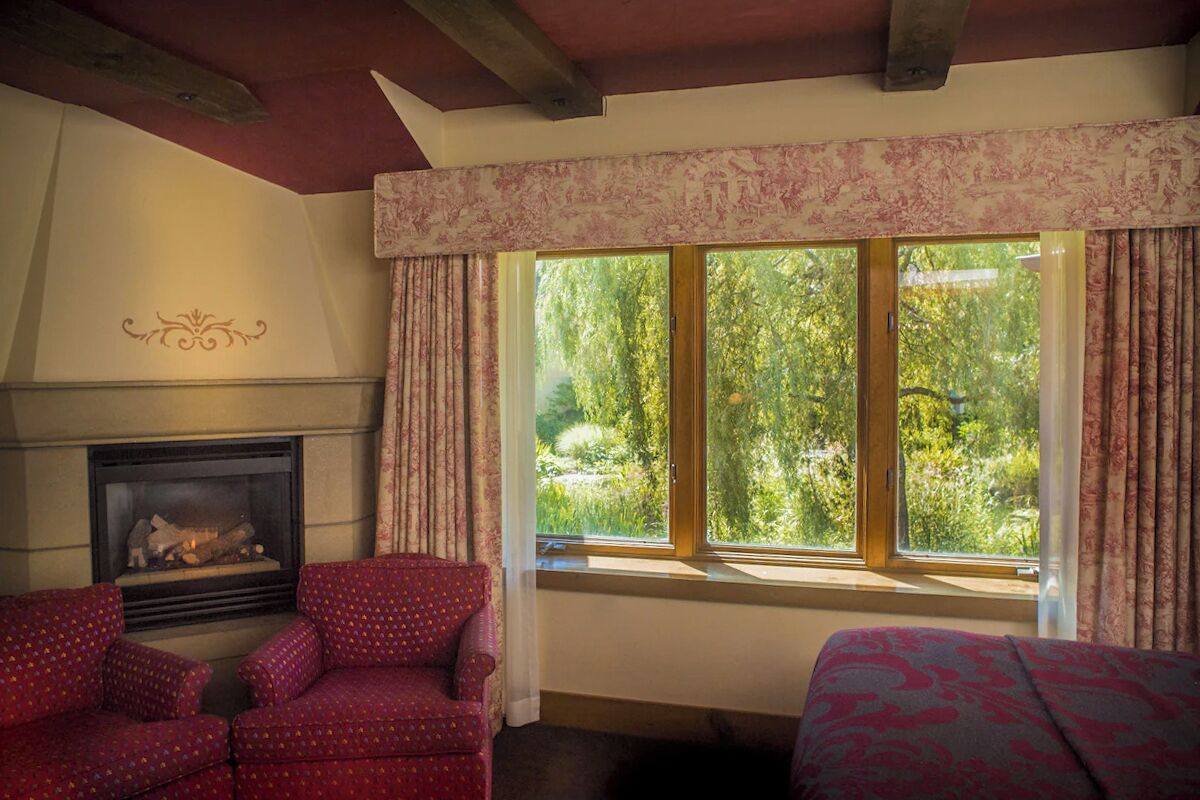 A room with a fireplace, two patterned armchairs, a bed, and a large window with curtains.