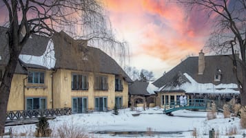 A snow-covered courtyard with a bridge, buildings with multiple windows, and a pond.