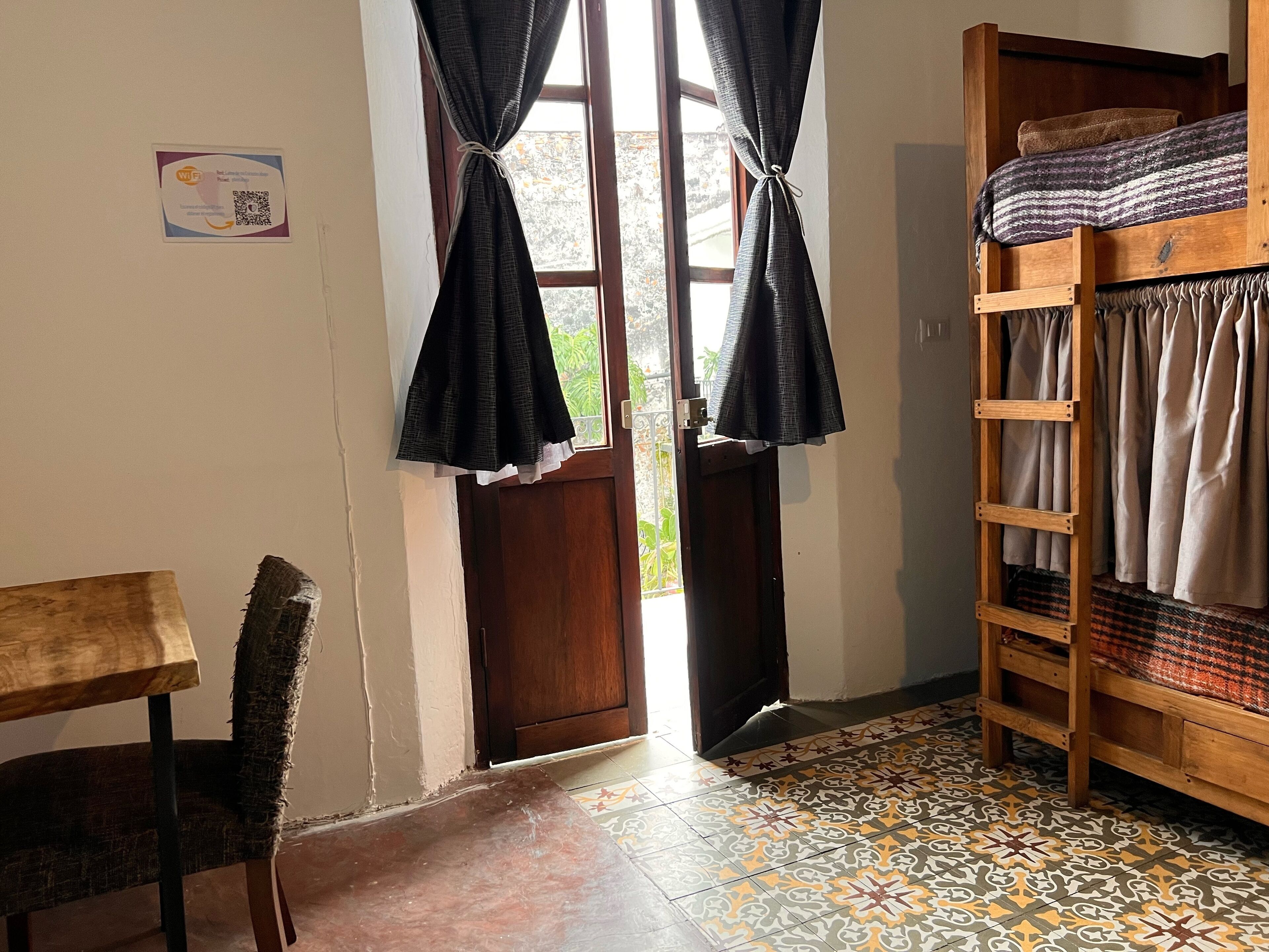A room with a wooden bunk bed, a small table, and a chair. There is a door leading outside and a window with curtains.