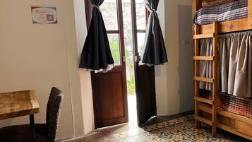 A room with a wooden bunk bed, a small table, and a chair. There is a door leading outside and a window with curtains.