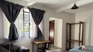 Habitación con dos camas, un escritorio y una silla. Tiene una ventana con cortinas, una puerta y un perchero.