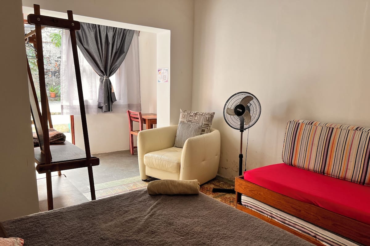 A living room with a beige sofa, a red sofa, a standing fan, a wooden chair, and a small table.