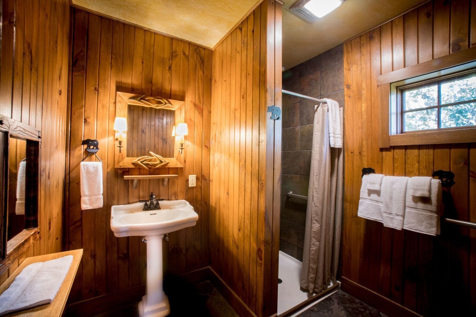 A wooden-paneled bathroom with a pedestal sink, a shower, and a window.