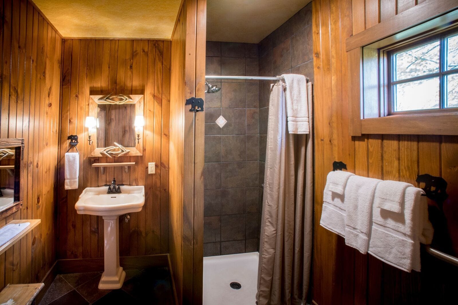 A wooden-paneled bathroom with a pedestal sink, a shower, and a window.