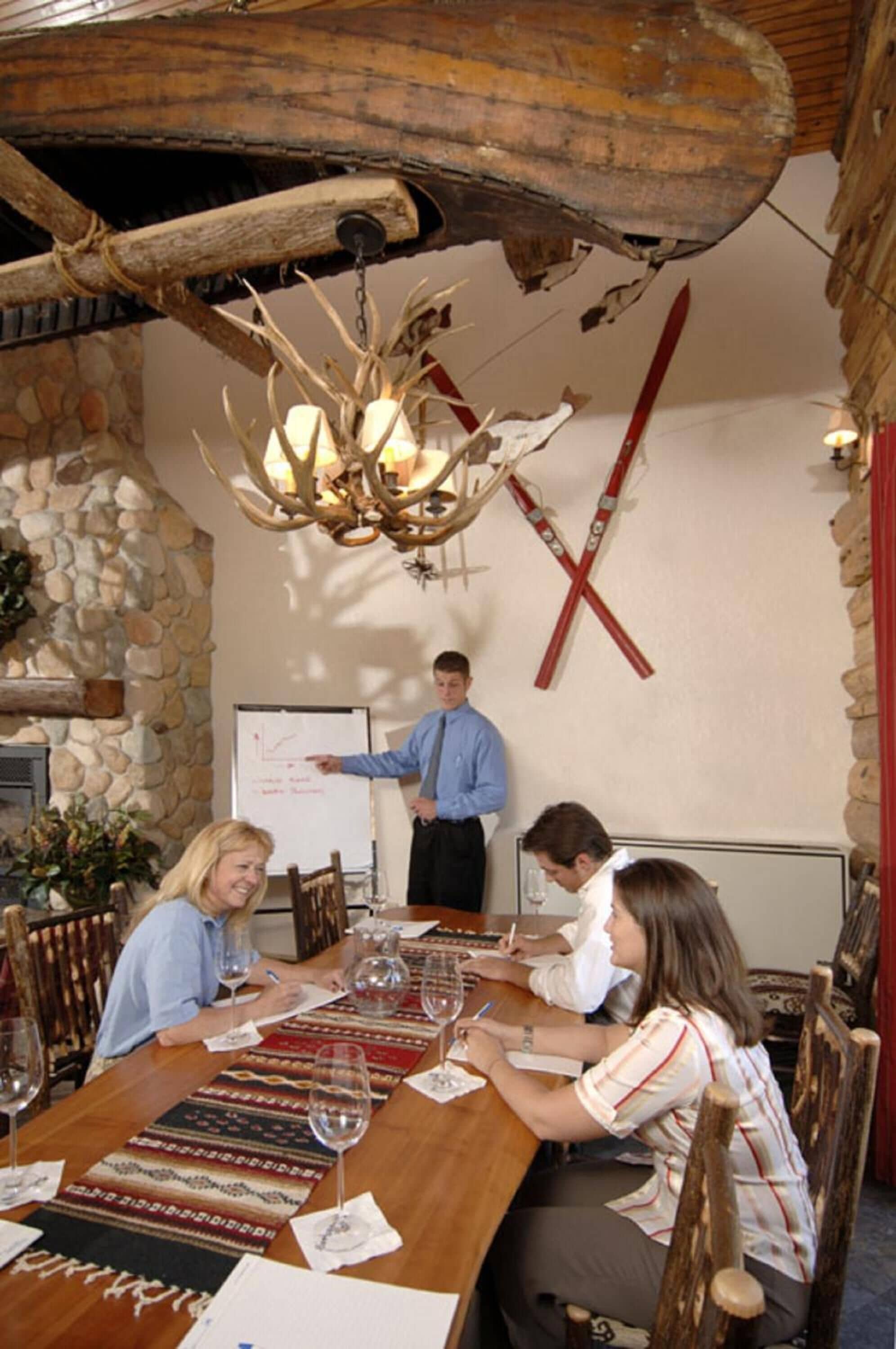 A wooden table with wine glasses, a whiteboard, and a person presenting.
