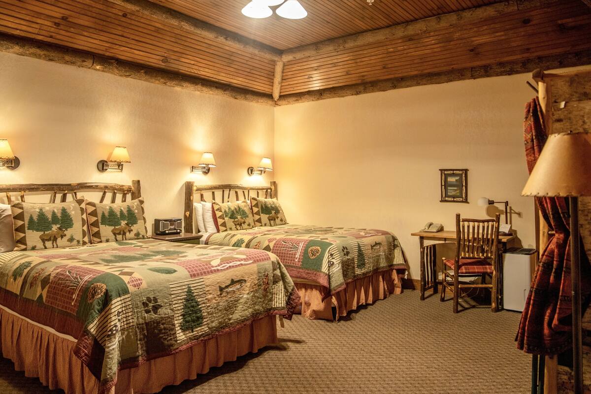 A room with two beds, a desk, and a chair. There are wall-mounted lamps and a framed picture on the wall.