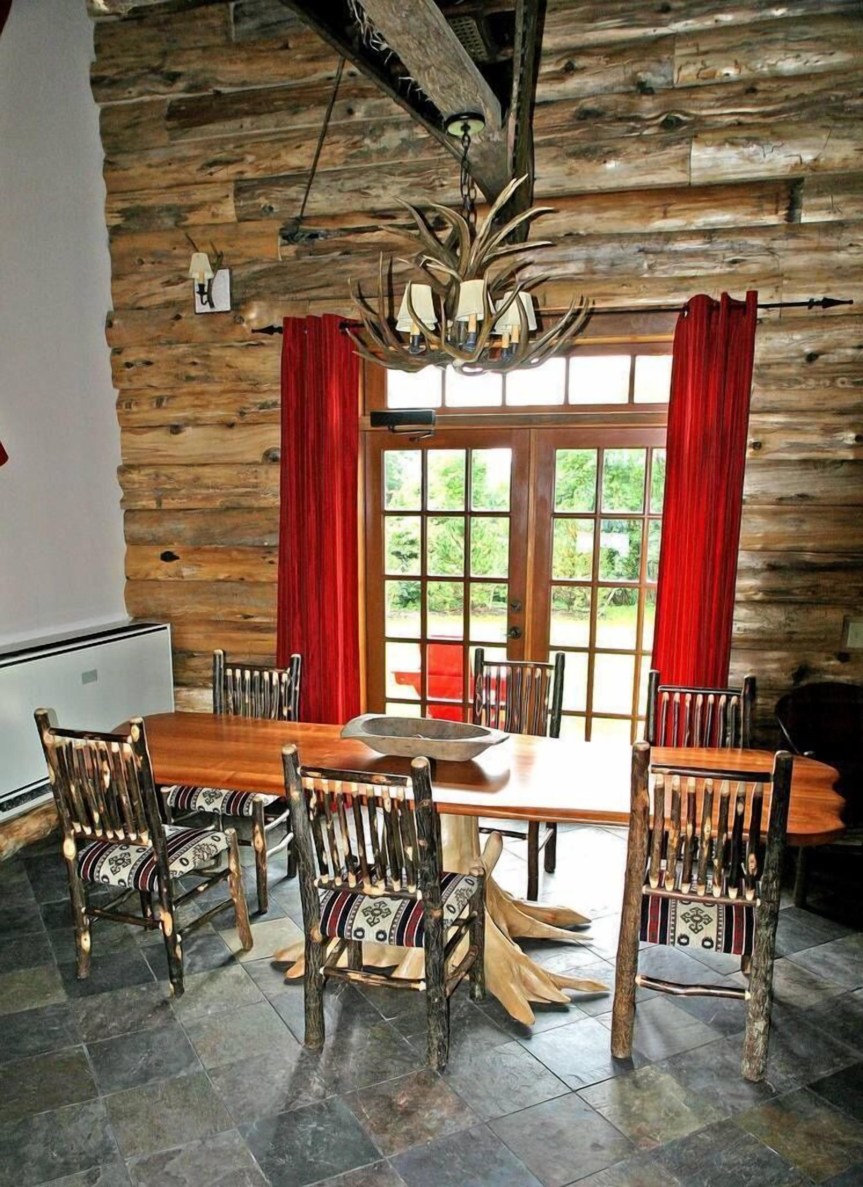Un comedor rústico con una mesa y sillas de madera, una lámpara de araña y vistas a través de un gran ventanal con cortinas rojas.