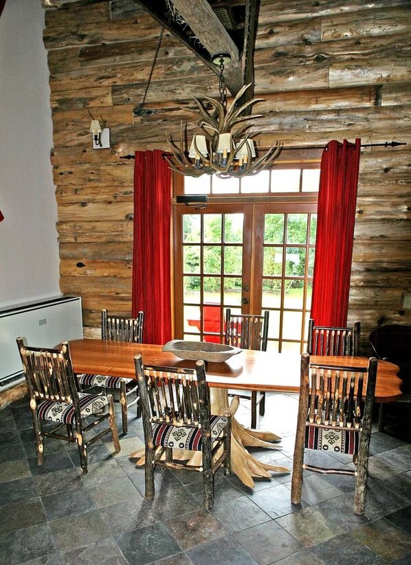Un comedor rústico con una mesa y sillas de madera, una lámpara de araña y vistas a través de un gran ventanal con cortinas rojas.