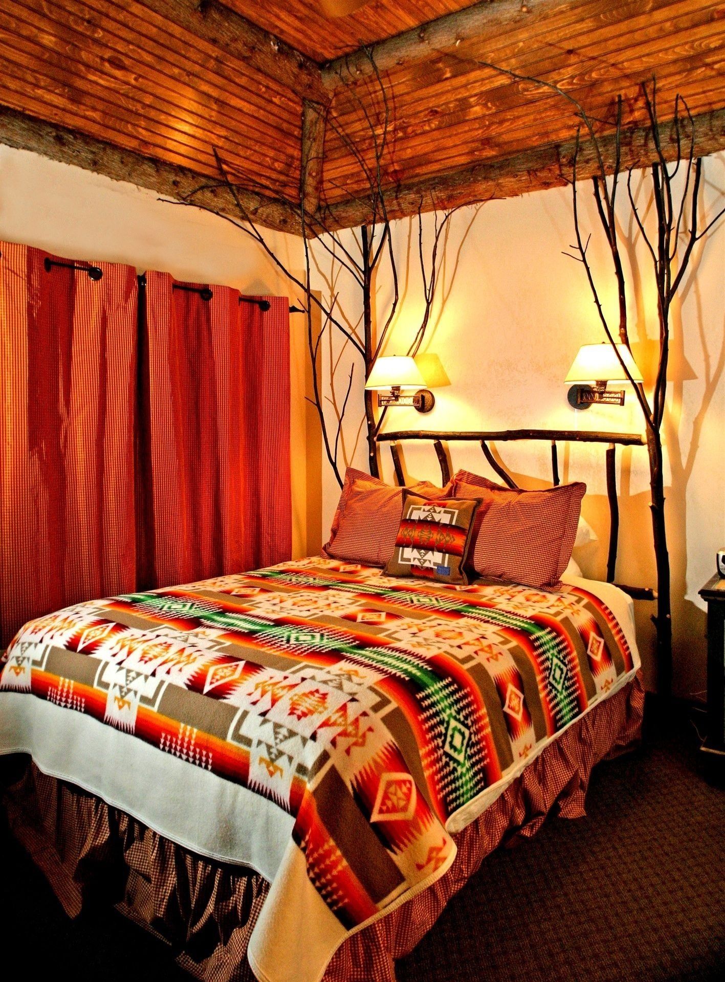 A bed with a patterned bedspread, a headboard with two wall-mounted lamps, and a red curtain.