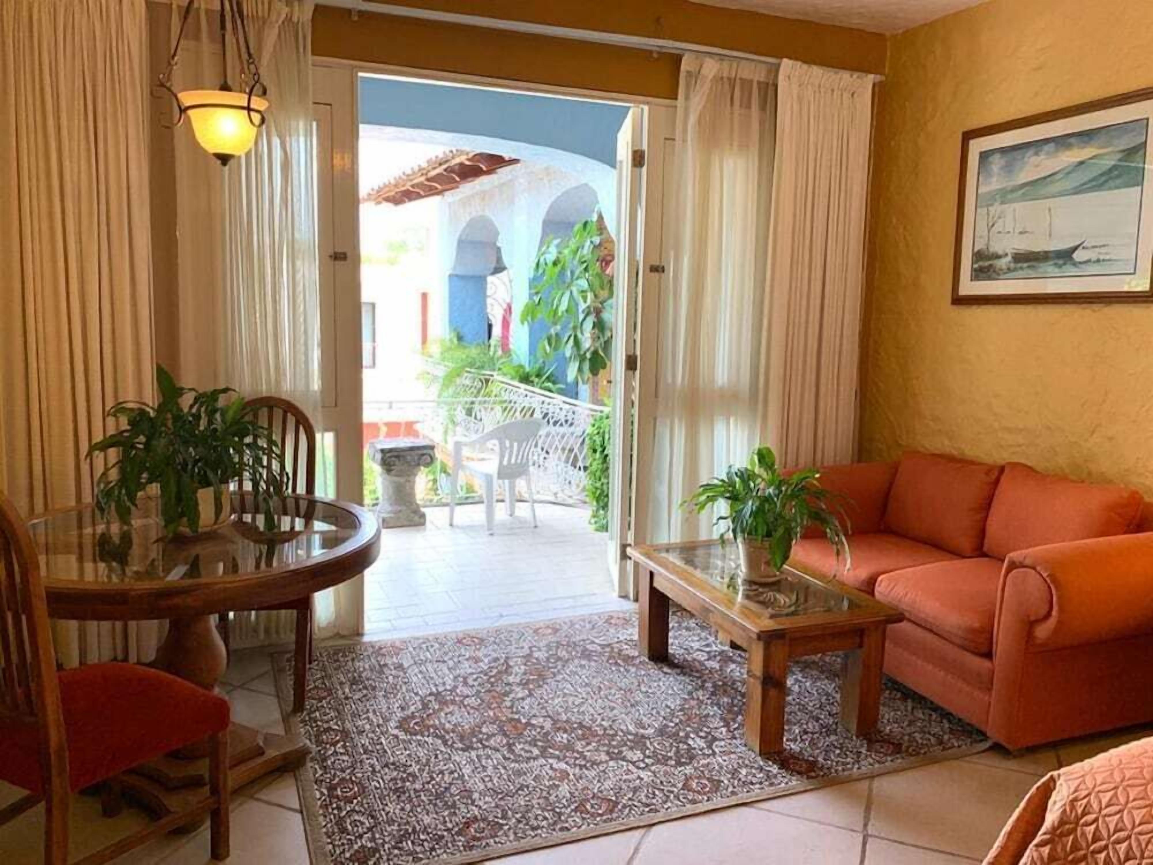 A living room with a sofa, a dining table with chairs, and a balcony with plants and a view of a building with arches.