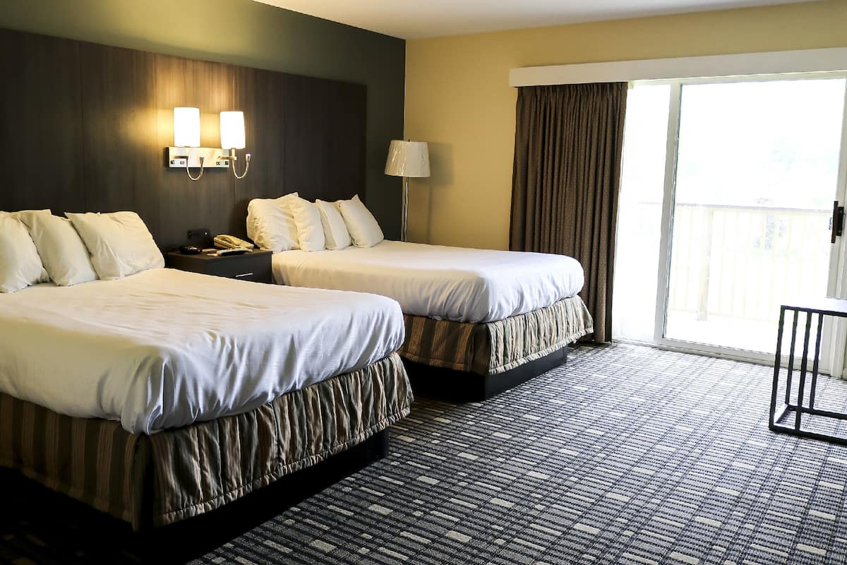 A hotel room with two beds, a nightstand, a lamp, and a view of the outdoors.