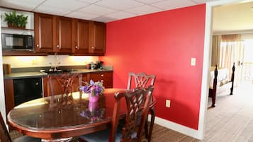 A dining area with a glass table and wooden chairs, a red accent wall, and a kitchen with wooden cabinets and a microwave.