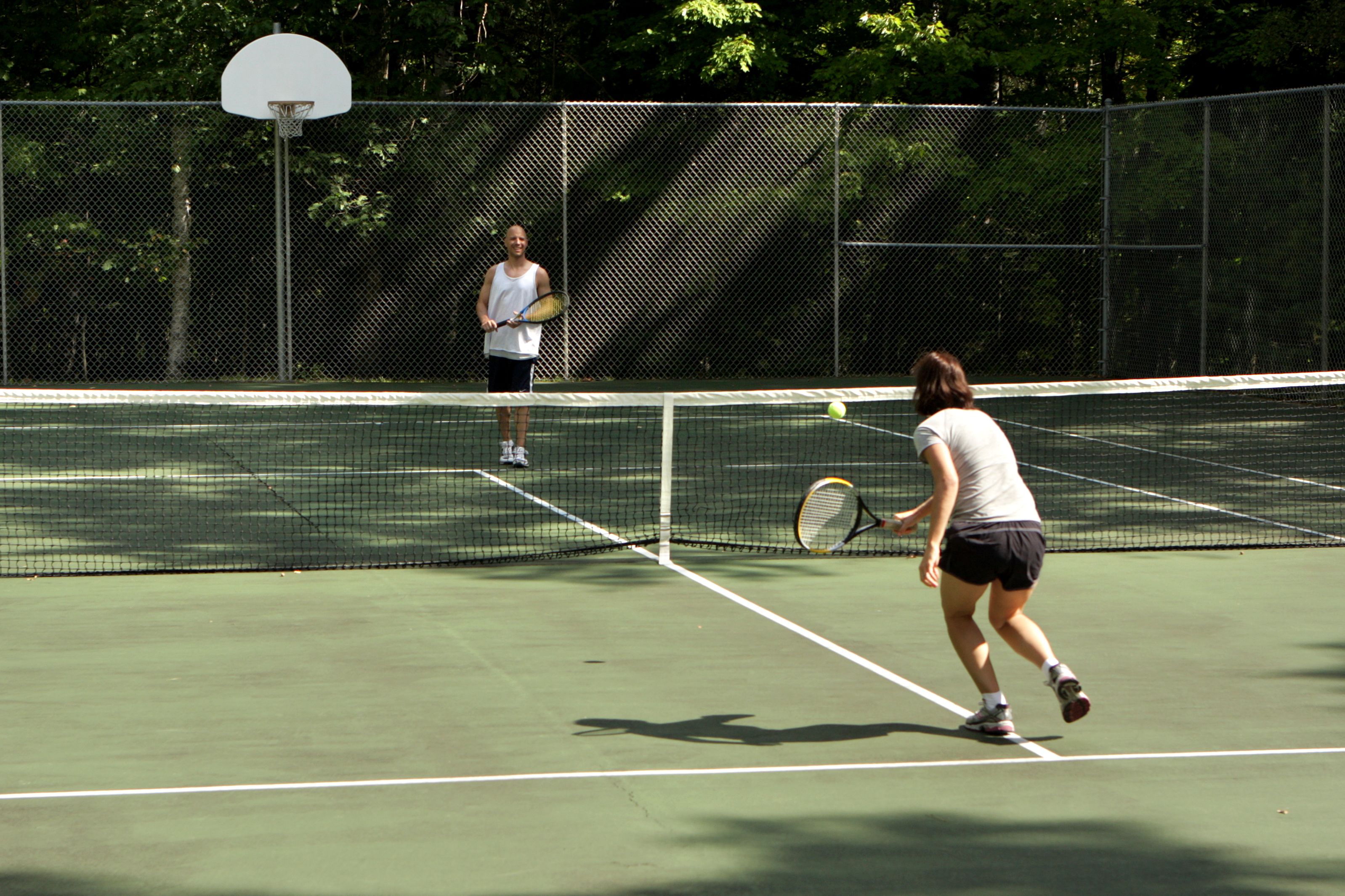 A tennis court with a net, two players, and a basketball hoop.