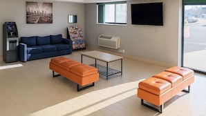 Ein modernes Wohnzimmer mit einem blauen Sofa, orangefarbenen Hocker, einem Couchtisch, einem Flachbildfernseher und einem Fenster mit Blick auf die Stadt.