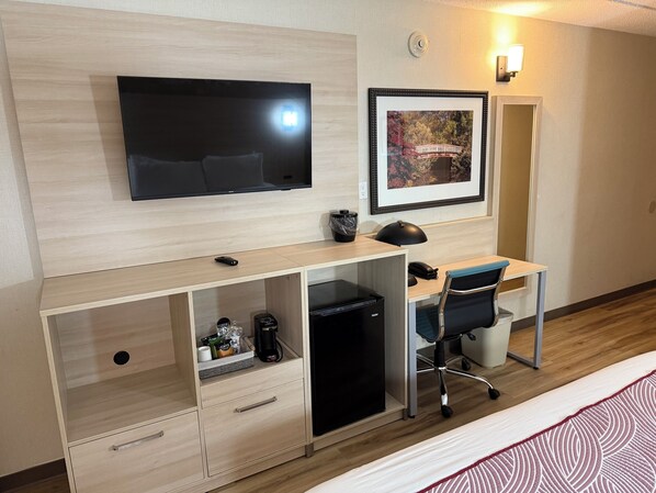 A hotel room with a flat-screen TV mounted on the wall, a desk with a chair, a coffee maker, and a small refrigerator.