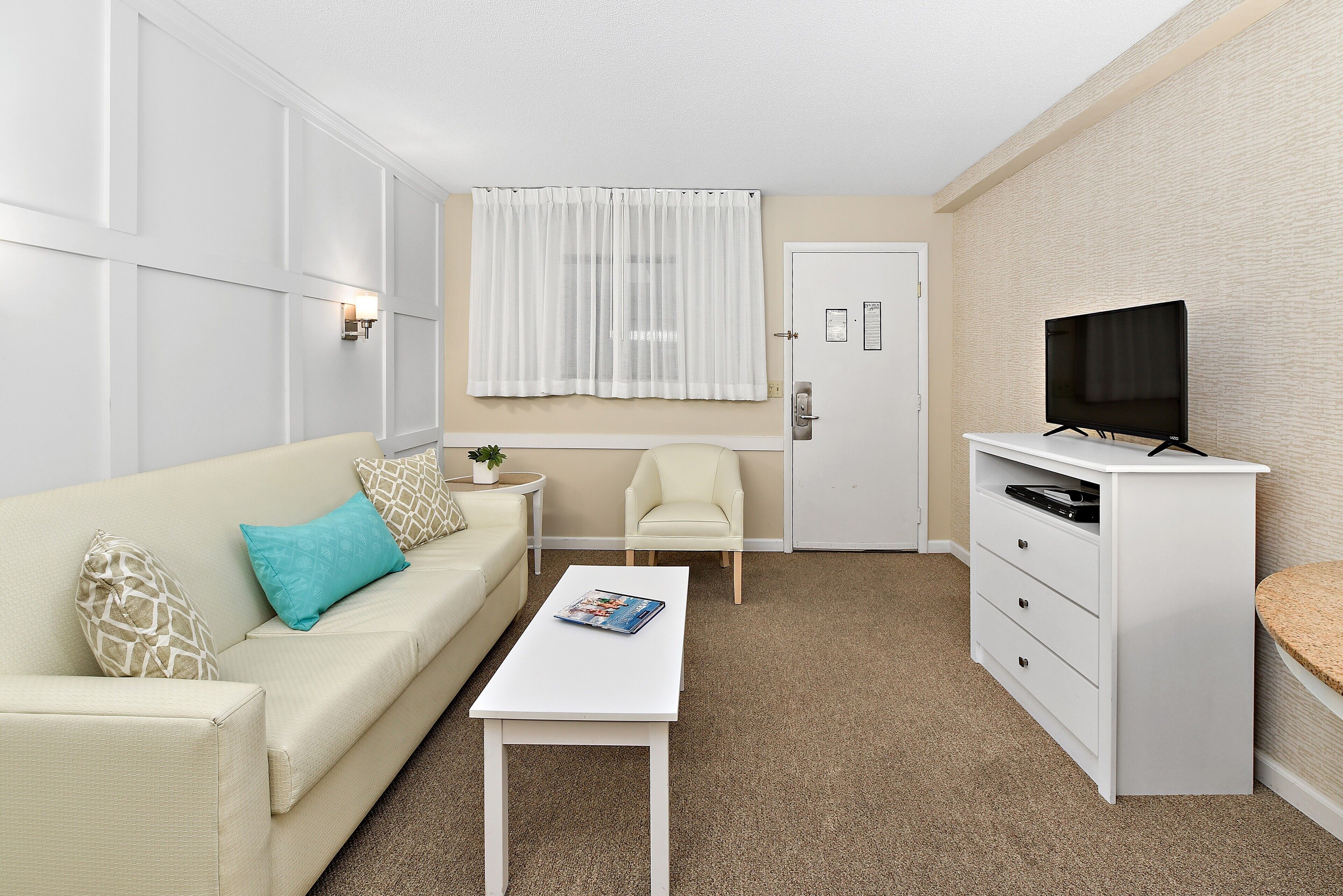 A compact living space with a white sofa, a small coffee table, a television on a cabinet, and a window with sheer curtains.