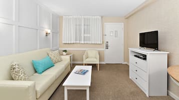 A compact living space with a white sofa, a small coffee table, a television on a cabinet, and a window with sheer curtains.
