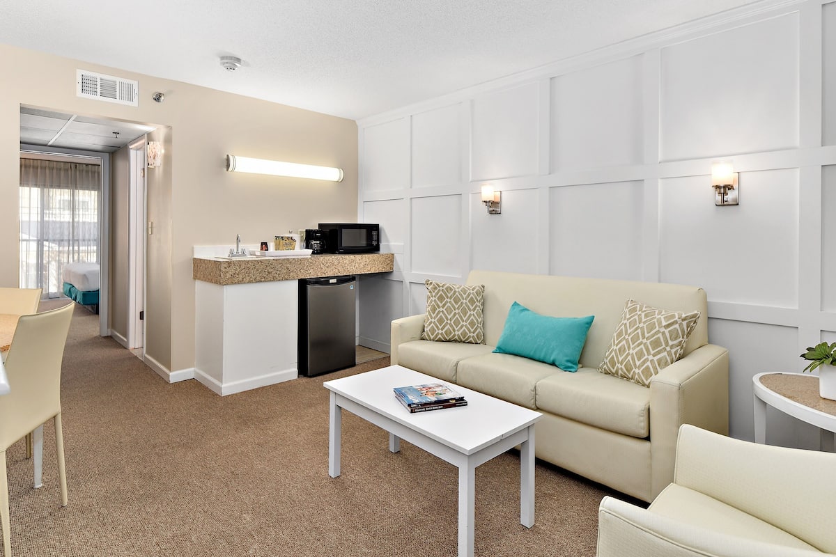 A hotel room with a kitchenette, a sofa with patterned cushions, a white coffee table, and a small side table with a plant.