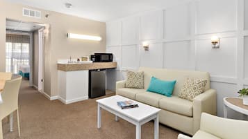 Et hotelværelse med et tekøkken, en sofa med mønstrede puder, et hvidt sofabord og et lille sengebord med en plante.