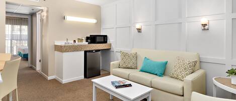 A hotel room with a kitchenette, a sofa with patterned cushions, a white coffee table, and a small side table with a plant.