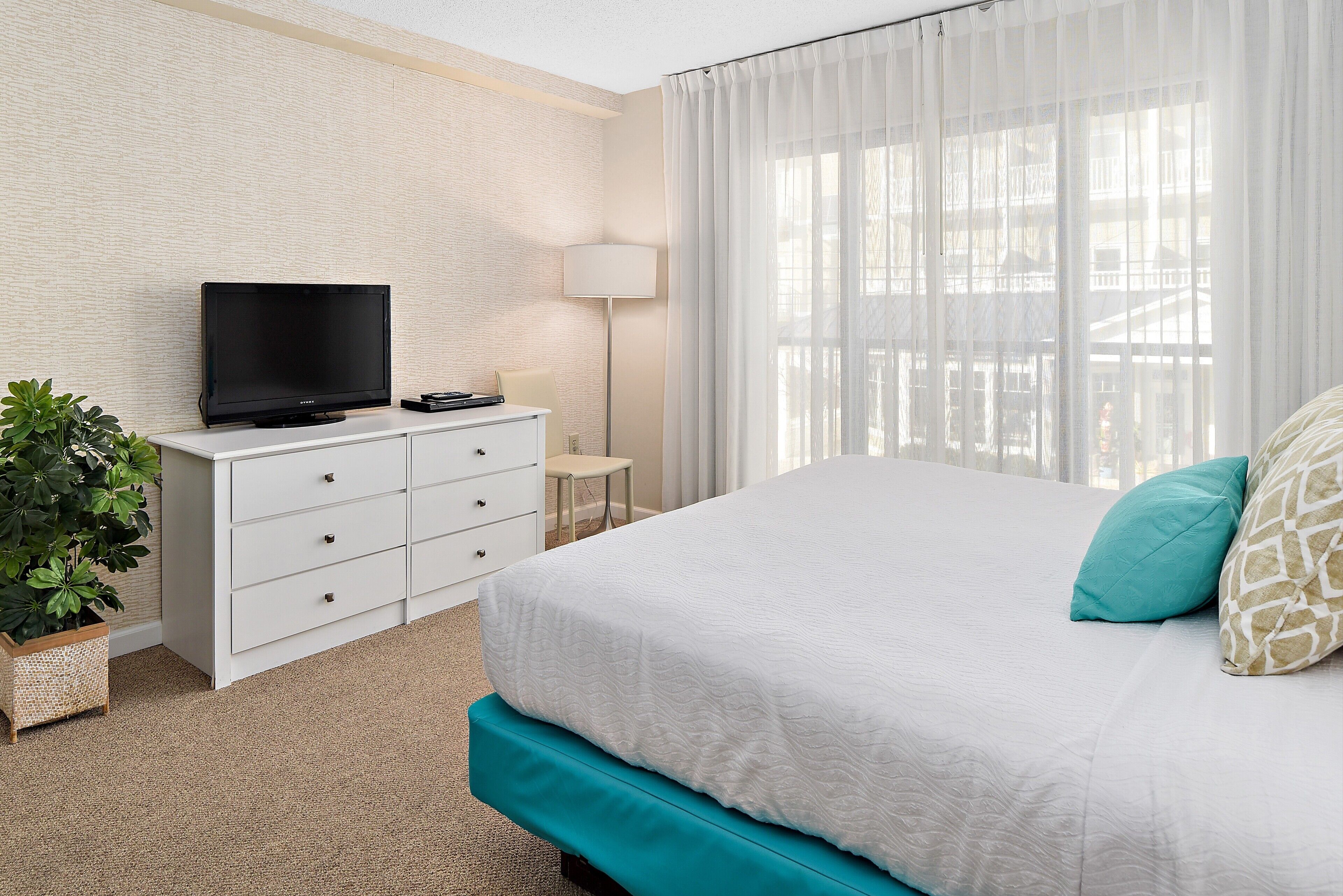 A hotel room with a bed, a TV on a white dresser, a lamp, a small table, and a plant.