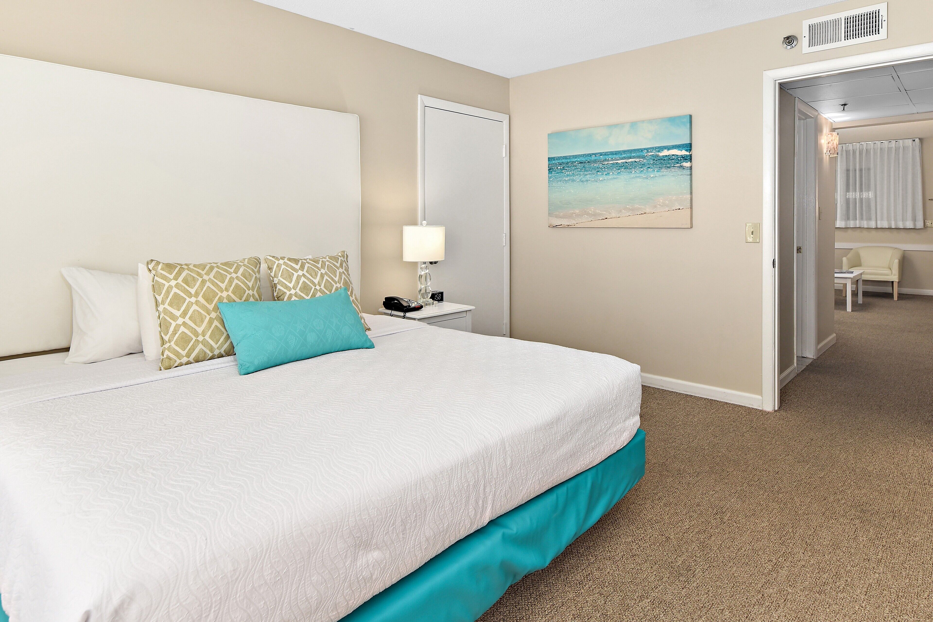 A hotel room with a large bed, a nightstand, a lamp, a painting of a beach, and a door leading to another room.