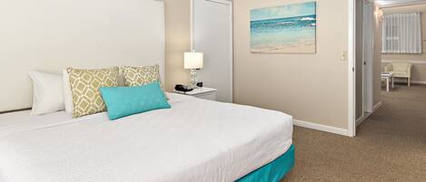 A hotel room with a large bed, a nightstand, a lamp, a painting of a beach, and a door leading to another room.