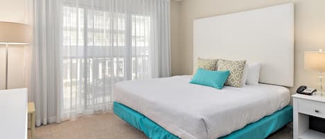 A hotel room with a large bed, a desk, a chair, and a window with sheer curtains.