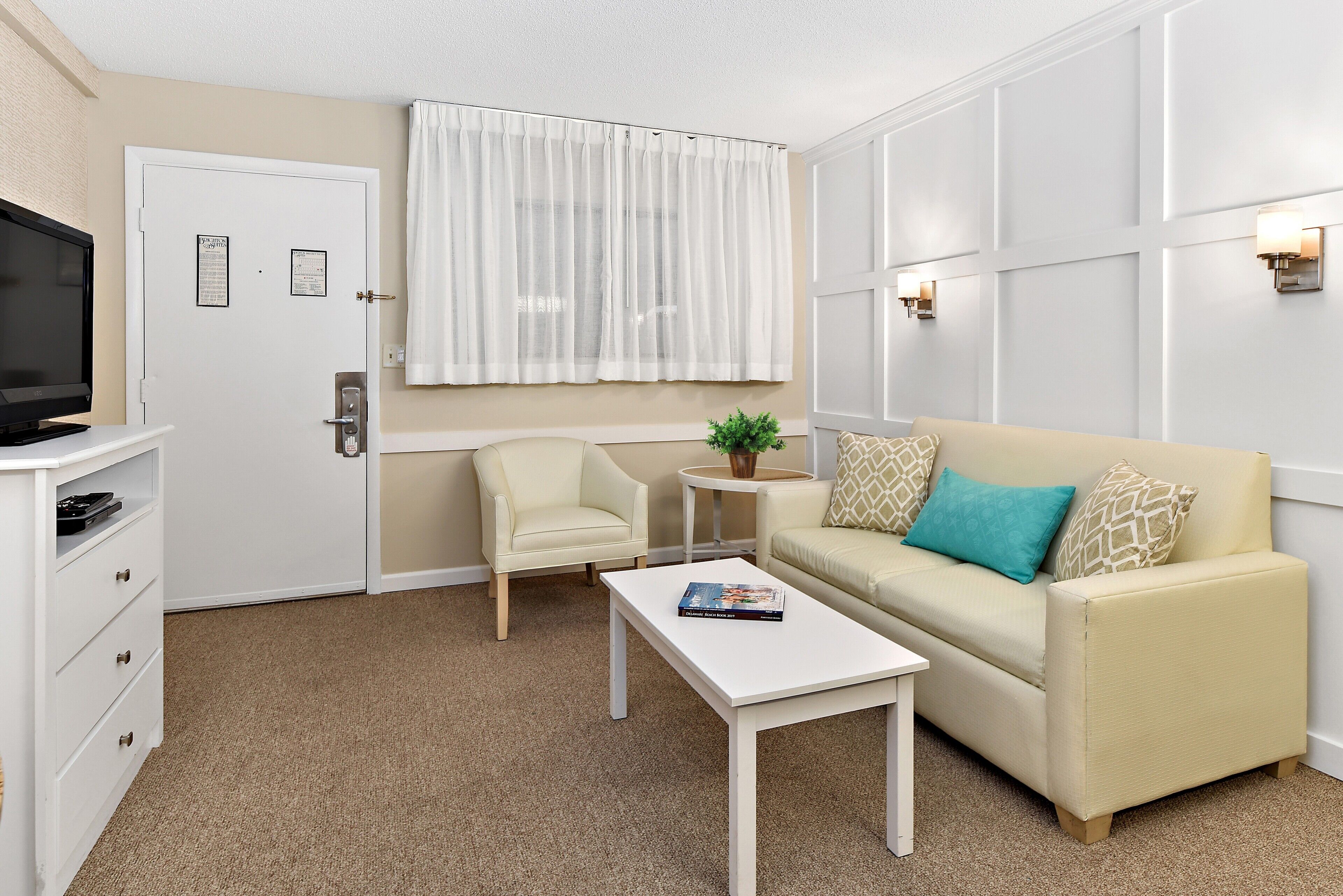 Una sala moderna con un sofá color beige, una mesa de centro blanca, una mesita auxiliar con una planta, un sillón blanco y un televisor sobre un mueble blanco.